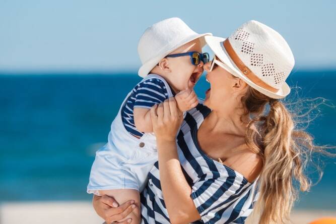 Mama i beba na plaži nose šešire i sunčane naočale za zaštitu od sunca